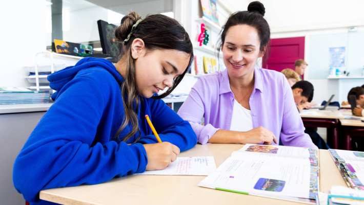 De lerares van de opleiding leraar basisonderwijs geeft persoonlijke uitleg aan leerling aan haar tafel