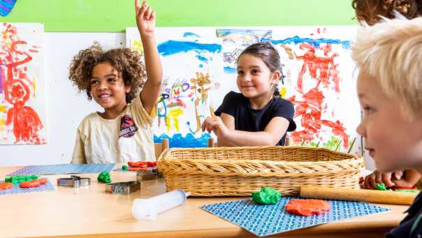 Drie kinderen zitten aan hun tafel in de klas, waarvan één kind zijn vinger opsteekt om een vraag te stellen. Foto voor de opleiding leraar basisonderwijs 