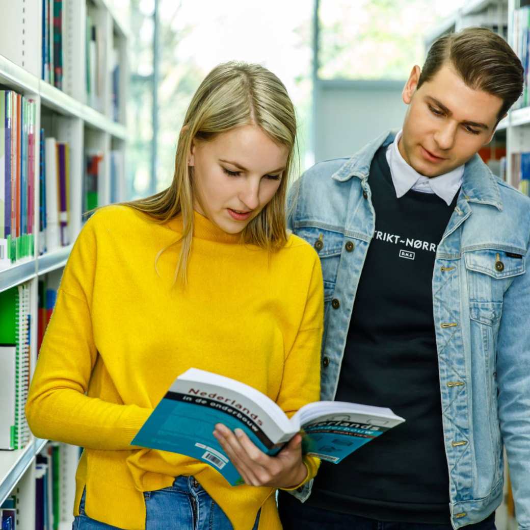 Twee studenten Leraar Nederlands bladeren door een boek in de mediatheek. 