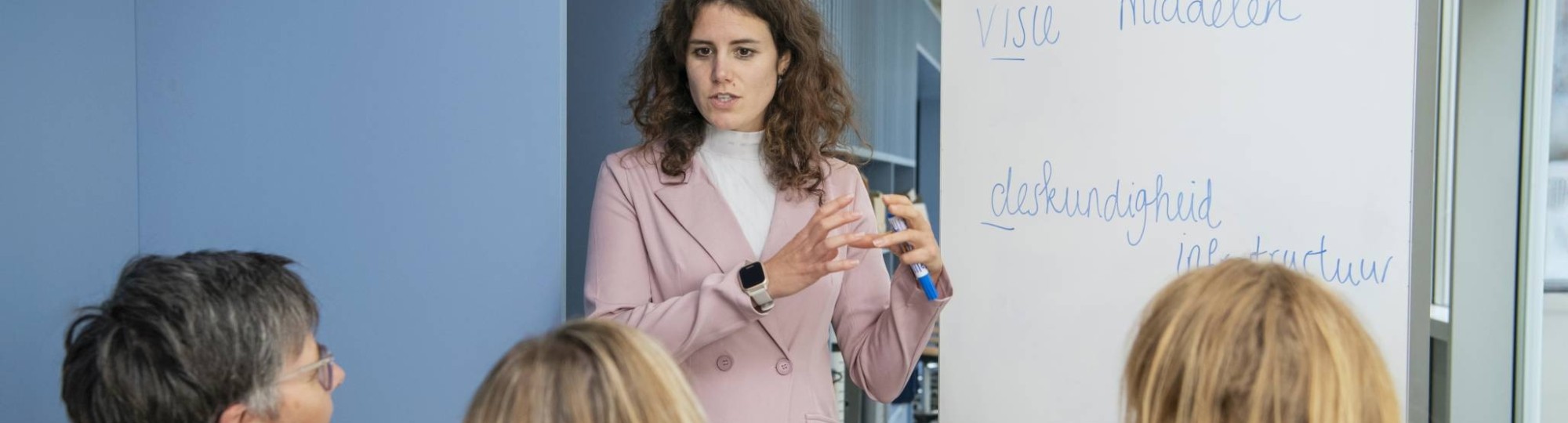 Jonge vrouwelijke student staat voor een whitebord en legt iets uit aan drie studenten die aandachtig luisteren.