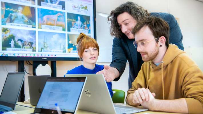 Lerarenopleiding Geschiedenis staat tussen twee studenten in en legt hen iets uit, terwijl iedereen op de laptop voor zich kijkt.