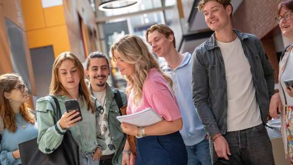 Groepje studenten staan bij elkaar en hebben plezier. Studenten kijkt op haar telefoon, ze staan in de gang. 