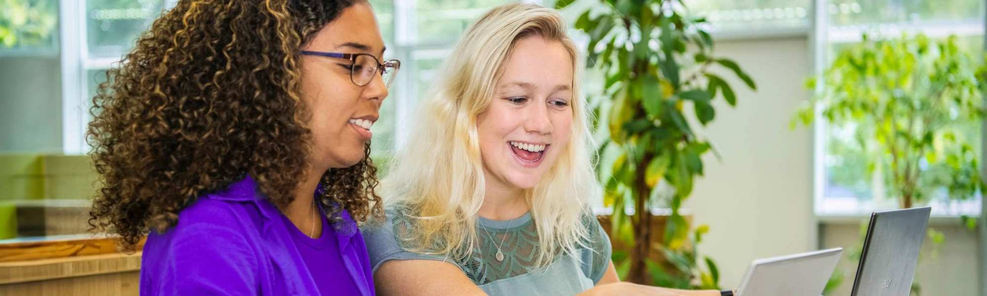 Twee studenten Leraar Wiskunde zitten naast elkaar met een laptop. De ene wijst lachend op het scherm iets aan en de ander kijkt lachend mee.
