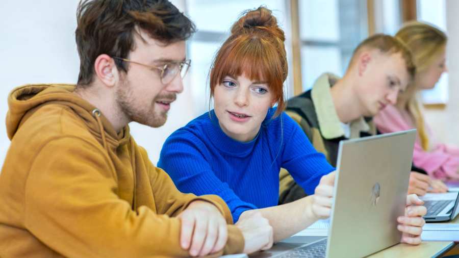 Twee studenten zitten naast elkaar en kijken samen aandachtig naar de laptop die voor hun staat.