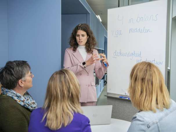 Jonge vrouwelijke student staat voor een whitebord en legt iets uit aan drie studenten die aandachtig luisteren.