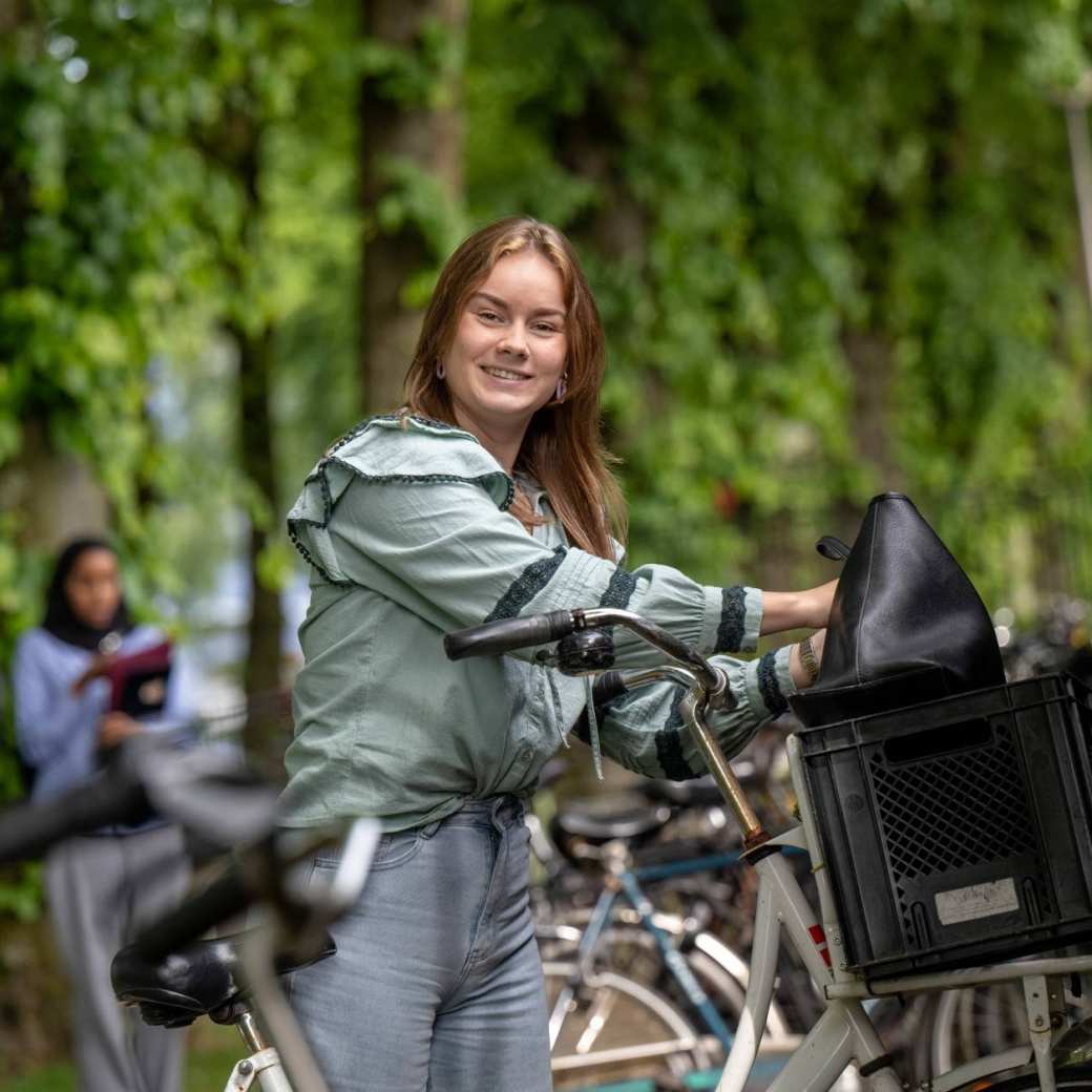 Student parkeert haar fiets in de fietsenstalling bij LvS10, kijkt in de camera, op campusterrein. 