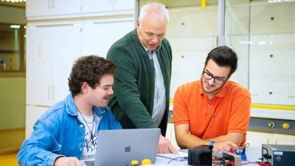 De leraar wijst twee studenten in een boekje wat aan.
