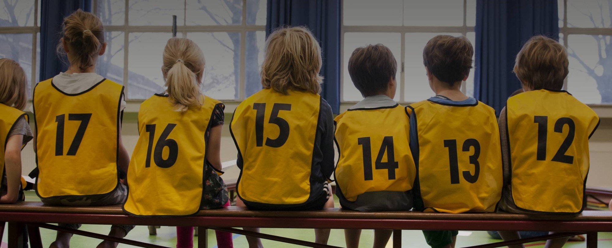 Kinderen in een gymzaal met hesjes aan op de rug gefotografeerd