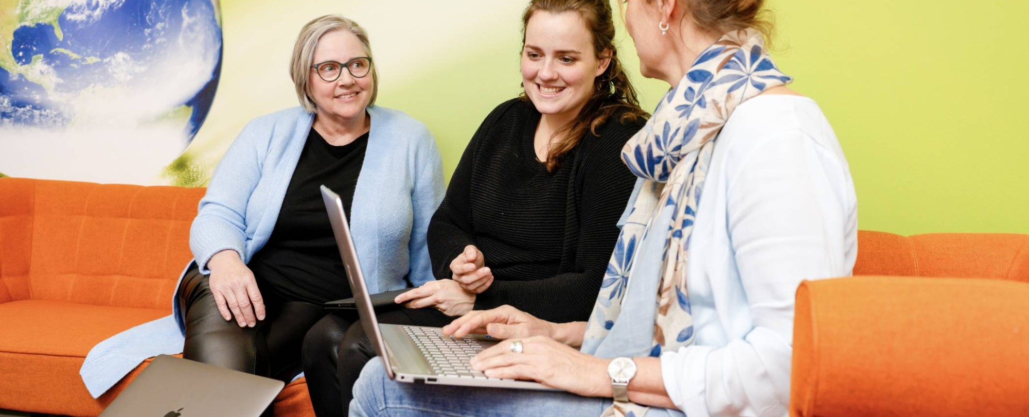 vrouw wijst iets aan op laptop, oranje banken. Wereldbol op achtergrond, tweede laptop op tafel.