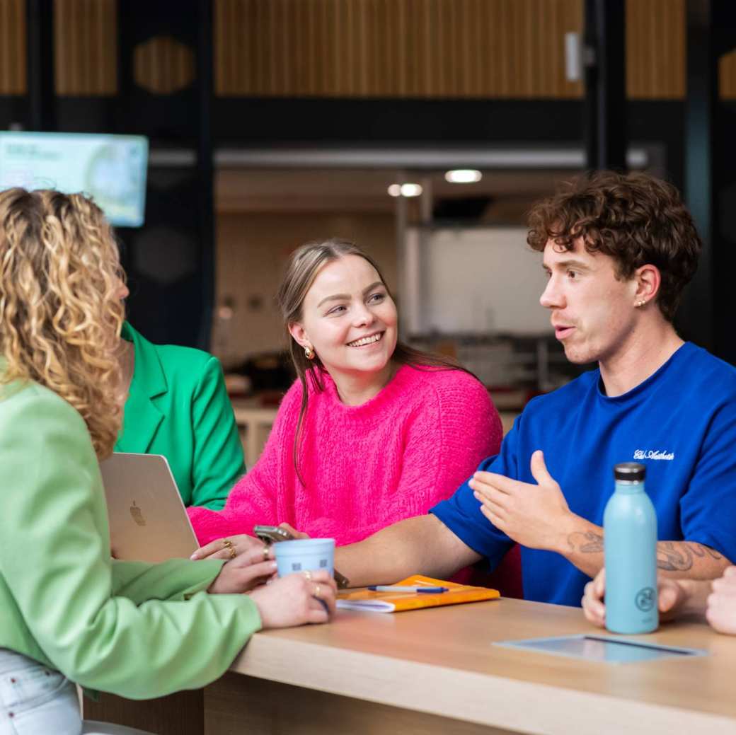 Groep studenten zit aan een lange tafel, lachen en kletsen samen.