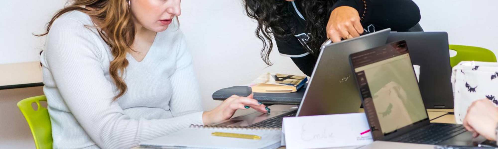 Twee studenten Leraar Engels kijken aandachtig naar de laptop en één student wijst naar het scherm.
