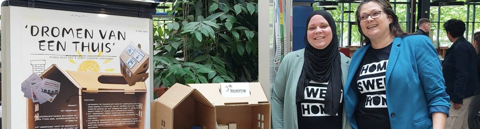 Studenten Joany en Marie staan voor een tafel waarop een soort poppenhuis te zien is. Op de poster naast hen staat informatie over een droomhuis.