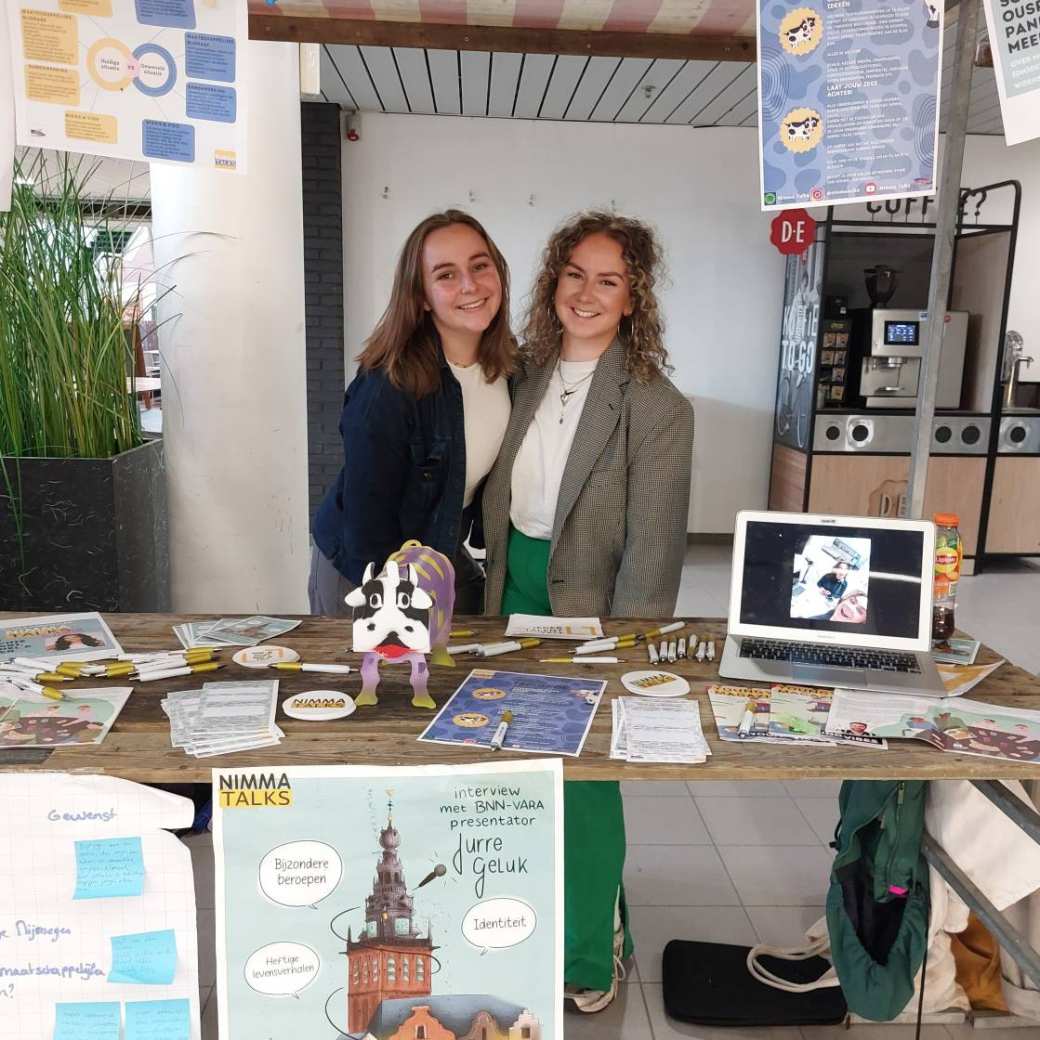 Links op de foto een vrouw met lichtbruin sluik haar en rechts blonde krullen en een grijze blazer. Ze staan in kraam volgehangen met posters.