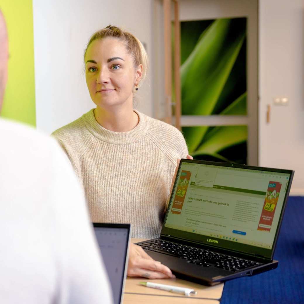 Vrouw laat laptop aan man zien, man van achterkant. Groene muur.