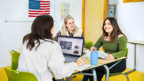 Drie studenten zitten in een Engels lokaal waar de Amerikaanse vlag op de achtergrond hangt