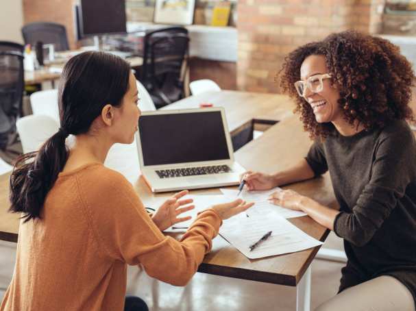 Twee vrouwen in gesprek in een kantoorsetting. Ze lachen en hebben een laptop en papieren.