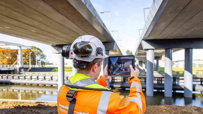 Man met veiligheidshelm en oranje werkjas fotografeert van onderen twee viaducten in aanbouw