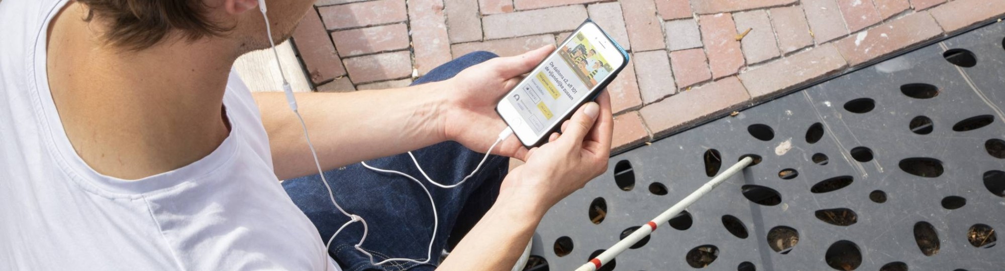 Jongen zit op een bankje en is met zijn mobiele telefoon bezig. Voor hem ligt zijn taststok.