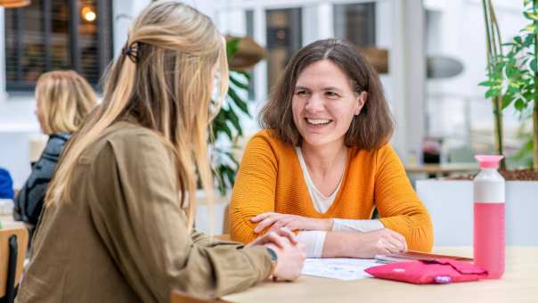 Vrouw in oranje trui lacht naar medestudent.