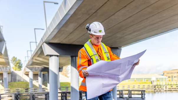 Man met veiligheidshelm en oranje werkjas checkt  de bouwtekening onder een viaduct in aanbouw.