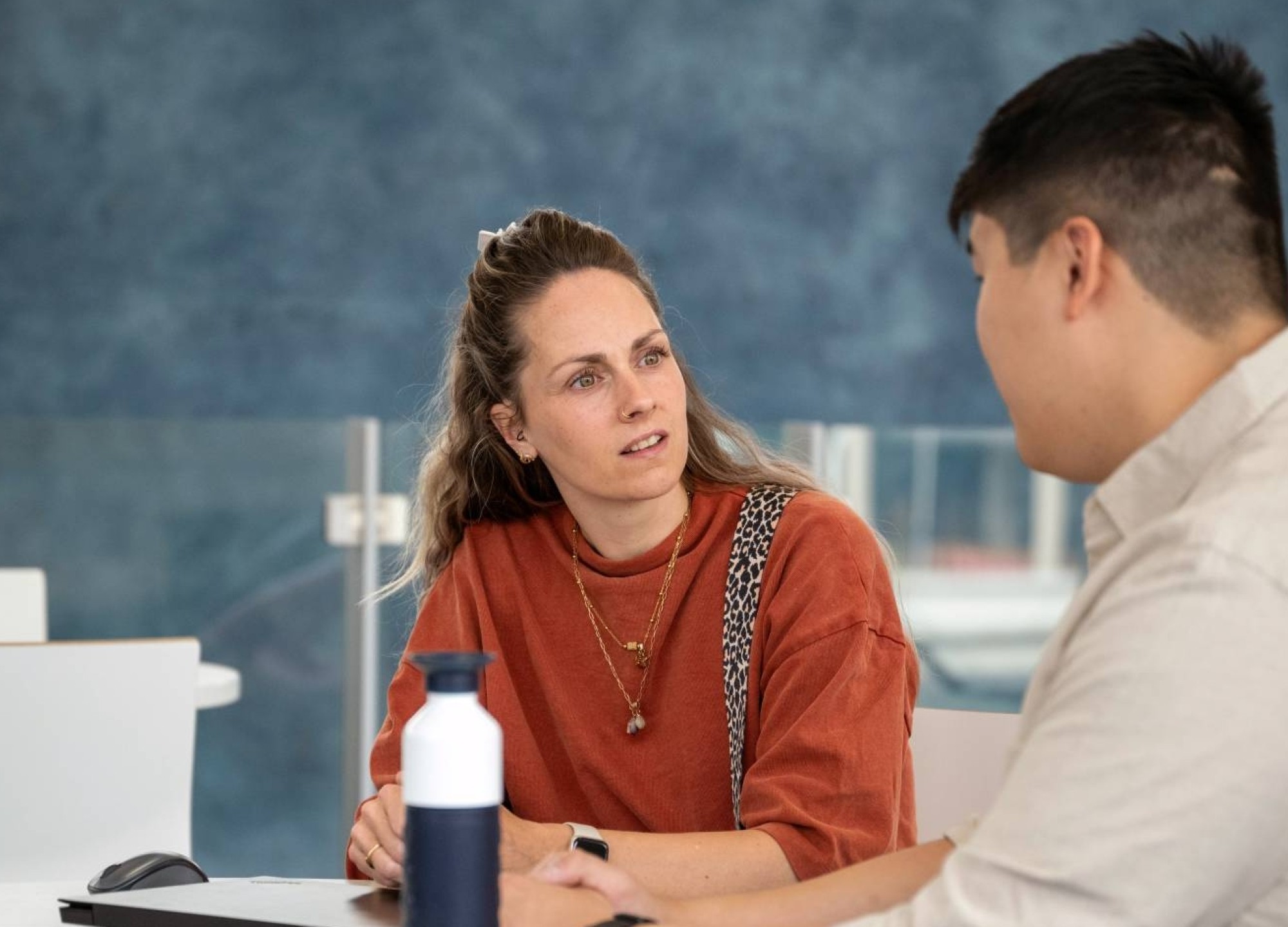 Docent in gesprek met student aan een tafel op podium in hal van gebouw R26, studentbegeleiding. 