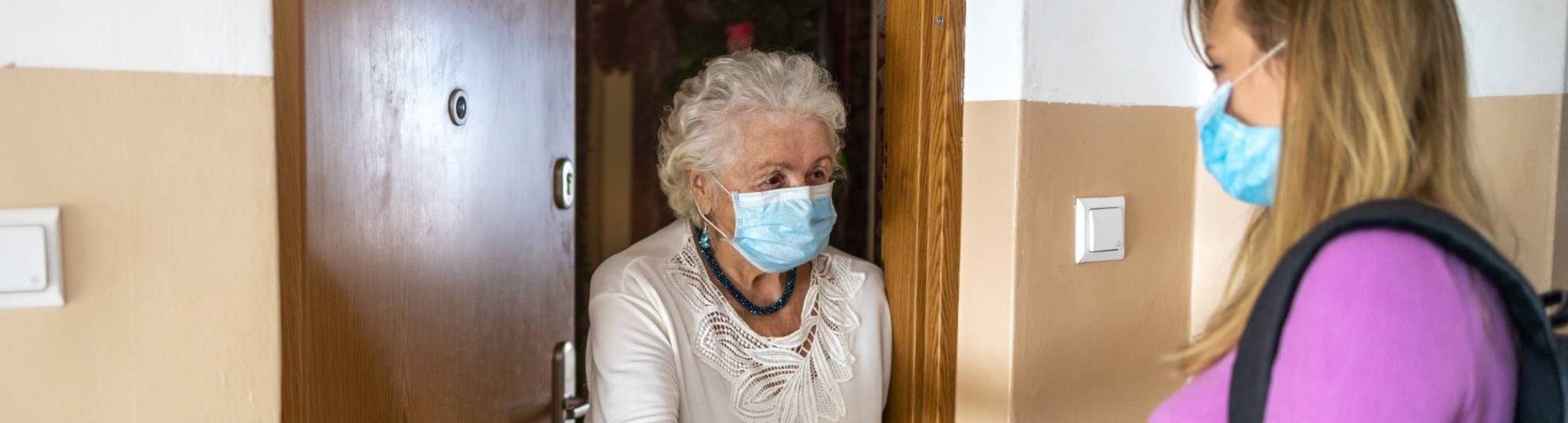 Een mantelzorgen komt tijdens coronatijd boodschappen brengen bij een oudere dame thuis. Met mondkapje.