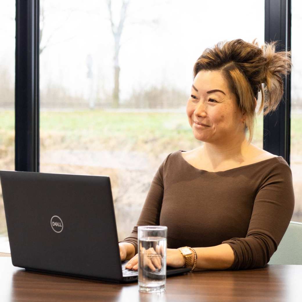 Dame poseert met een glimlach, achter een laptop. Een glas water staat naast haar op het bureau.
