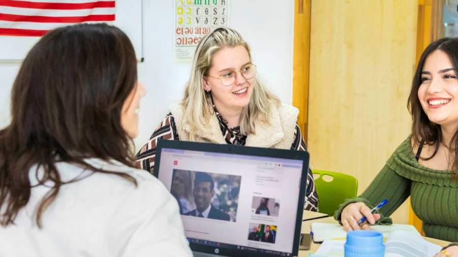 Drie studenten zitten in een Engels lokaal waar de Amerikaanse vlag op de achtergrond hangt