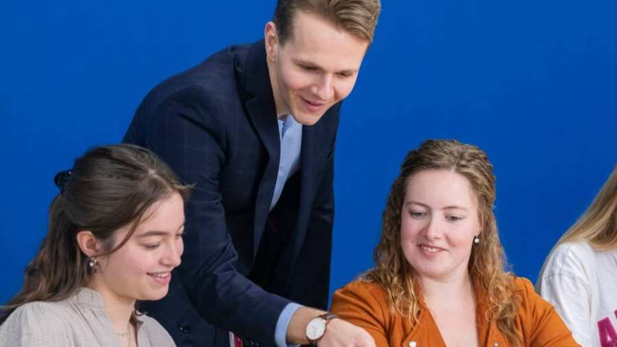 Docent in donkerblauw pak kijkt mee op de laptop bij twee van zijn studenten.
