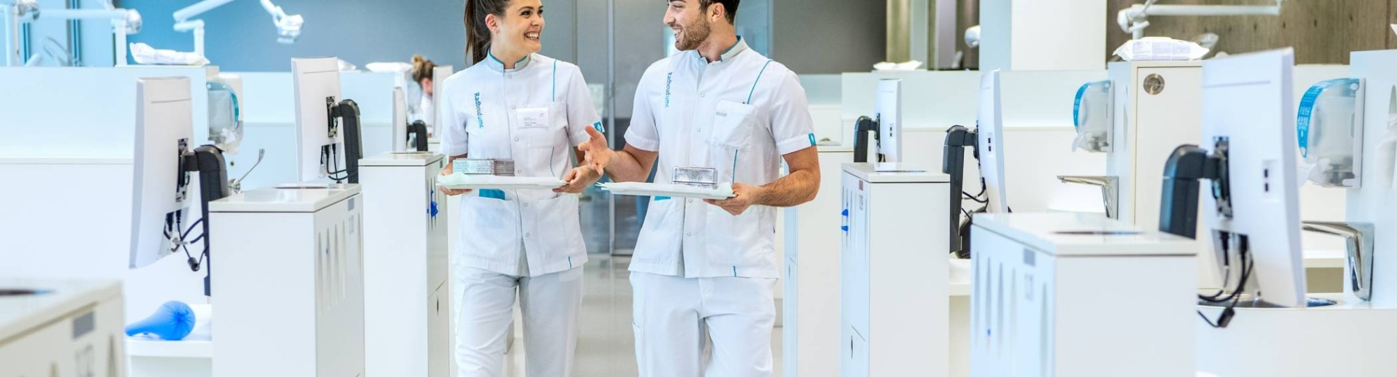 2 studenten kijken elkaar aan en lopen door een ruimte met veel behandelstoelen.