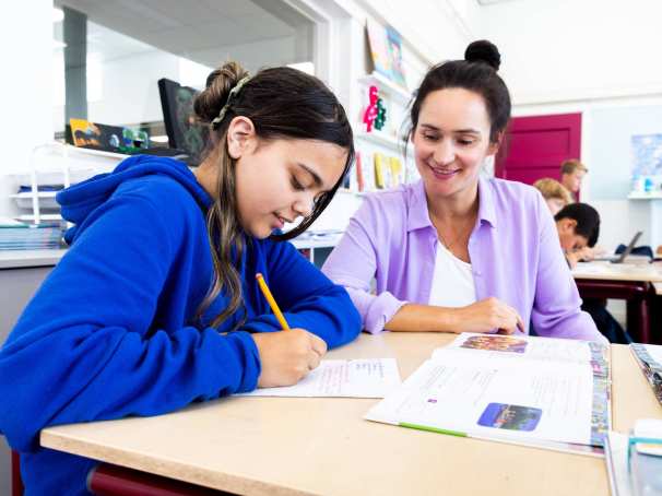 De lerares van de opleiding leraar basisonderwijs geeft persoonlijke uitleg aan leerling aan haar tafel