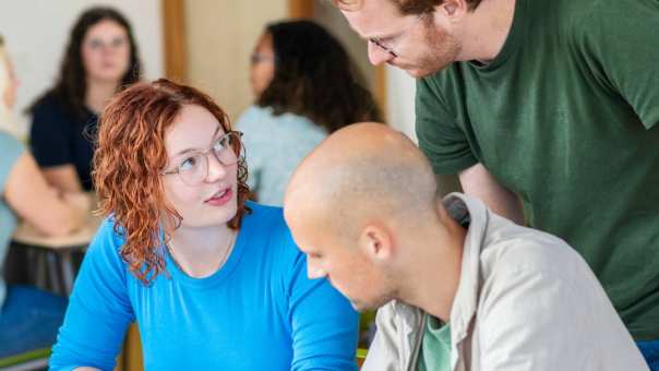 Leraar Wiskunde kijkt mee met zijn twee studenten, van wie eentje iets aanwijst in het schrift
