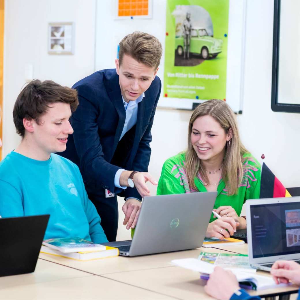 Docent Duits in pak geeft uitleg aan drie van zijn studenten, terwijl hij naar de laptop kijkt.
