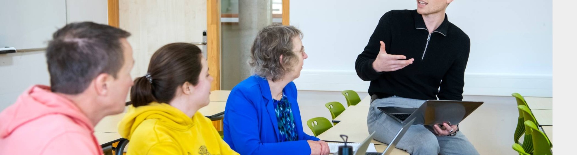 Foto Master Academie Educatie. 4 masterstudenten zitten in een lokaal. 1 van hen zit op een bureau en legt met een laptop in z'n hand wat uit aan de rest. 