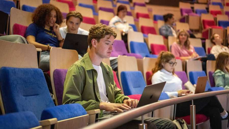 In collegezaal zit mannelijke student op laptop en maakt aantekeningen. 