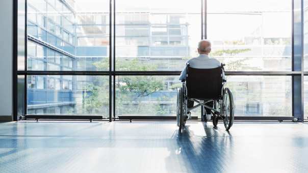 Een oudere man in een rolstoel staat alleen in een gang in het ziekenhuis en kijkt uit het raam.
