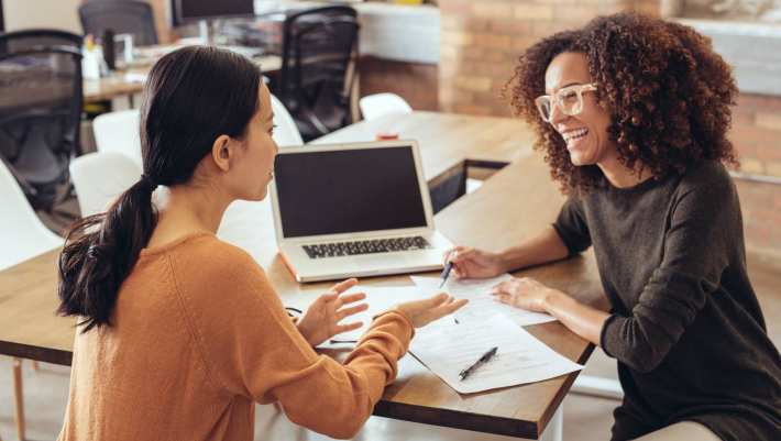 Twee vrouwen in gesprek in een kantoorsetting. Ze lachen en hebben een laptop en papieren.