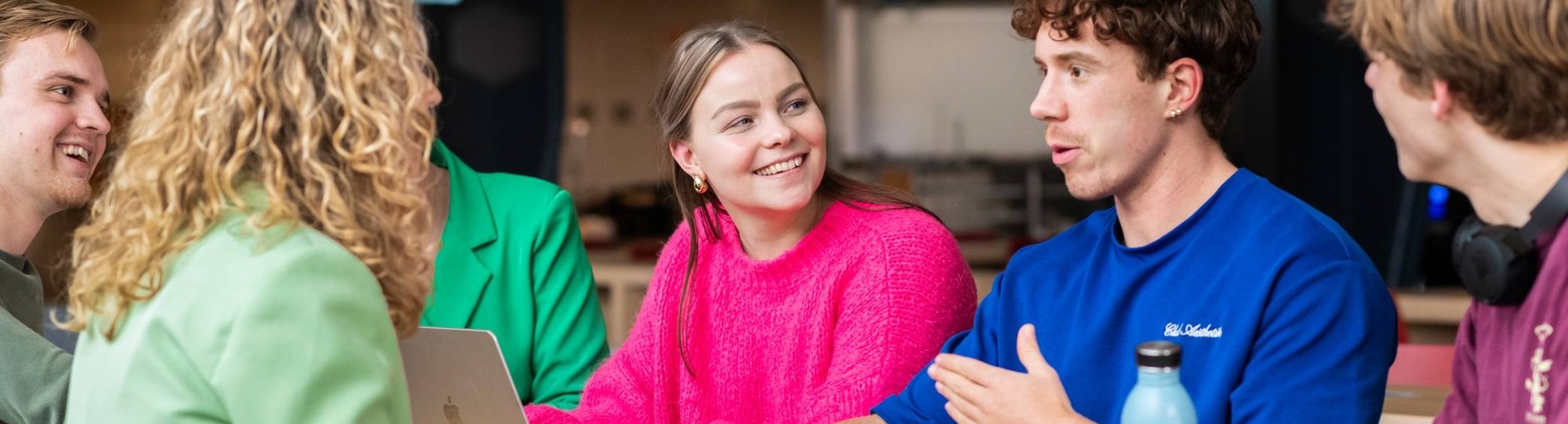 Groep studenten zit aan een lange tafel, lachen en kletsen samen.