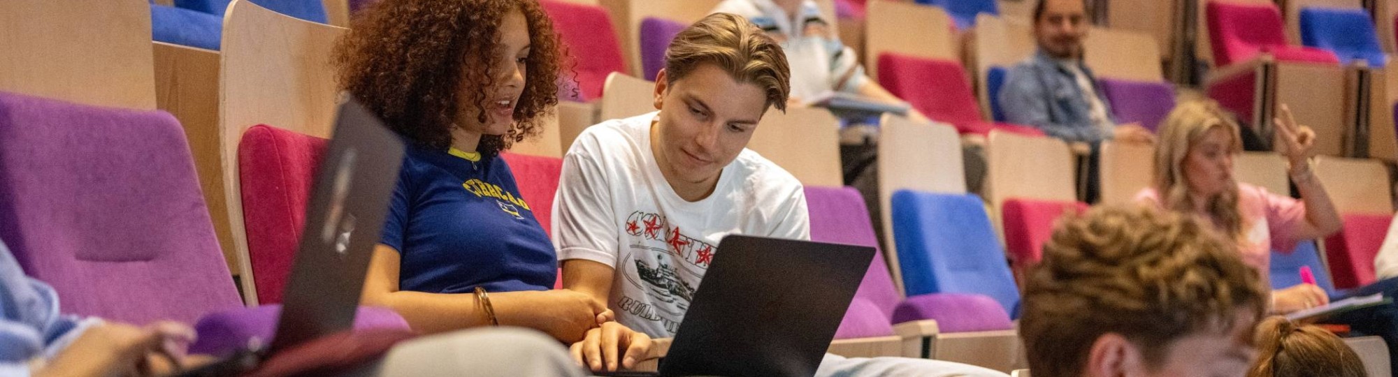 Twee studenten kijken samen op de laptop en overleggen in de collegezaal.