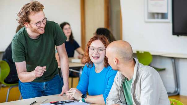 Leraar Wiskunde luistert lachend naar zijn twee studenten die iets zeggen tegen hem
