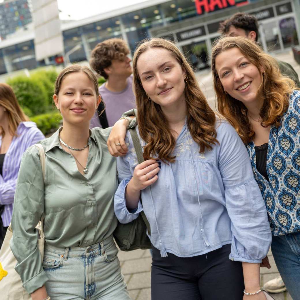 Studenten staan met vriendengroep buiten bij de ingang van gebouw R26, kijken in de camera.