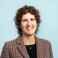 Portrait headshot. The picture is taken in front of a blue background and the subject is wearing a brown grid-patterned knit blazer with a black shirt underneath. Her hair is curly and brown and her eyes are hazel. She is facing the camera, with shoulders slightly towards the right. 