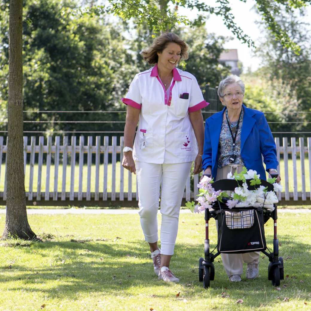 Zorgverlener wandelt met oudere dame in de tuin van een verpleeghuis.