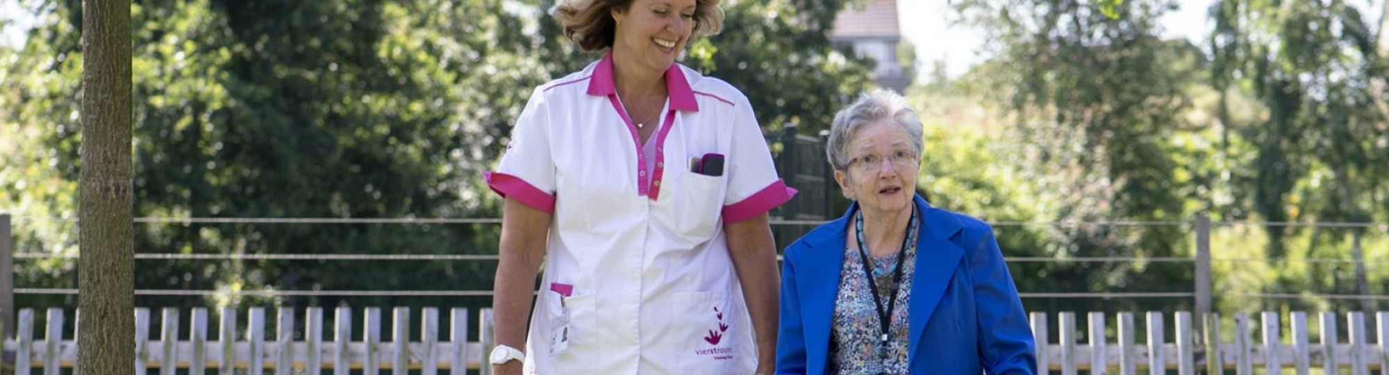 Zorgverlener wandelt met oudere dame in de tuin van een verpleeghuis.