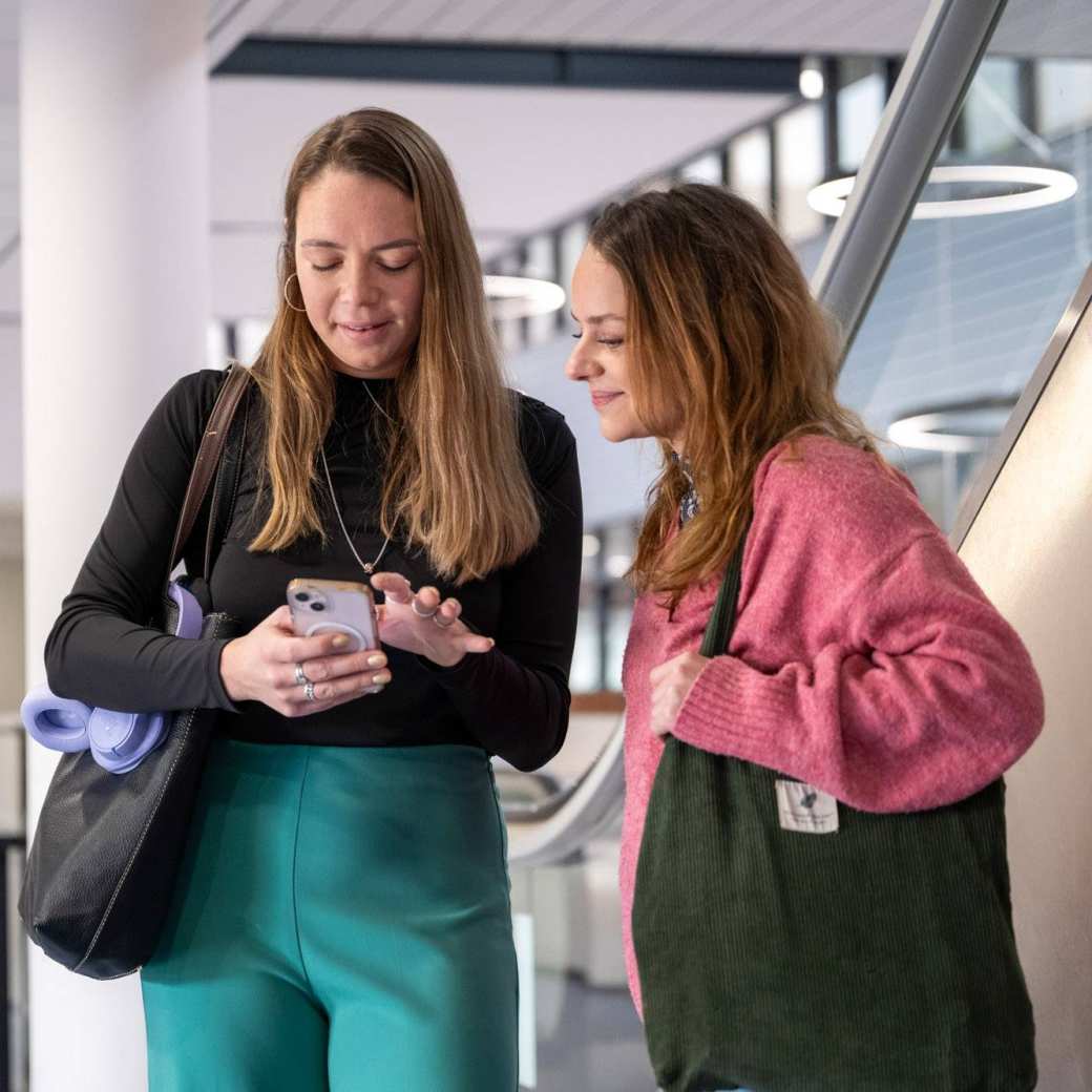 2 duale studenten kijken op een mobieltje in de hal.