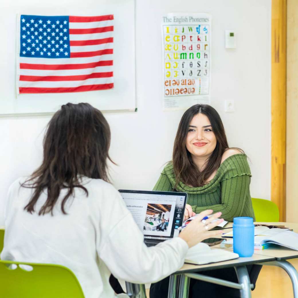 Studente met zwart haar en groene strakke top lacht met haar lippen op elkaar naar de persoon die voor haar zit. De Amerikaanse vlag hangt op de achtergrond.