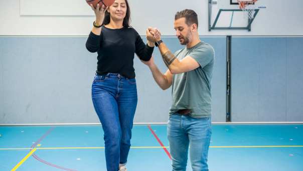 Twee studenten waarbij één student de andere student blind en met bal in de hand over een balk begeleidt