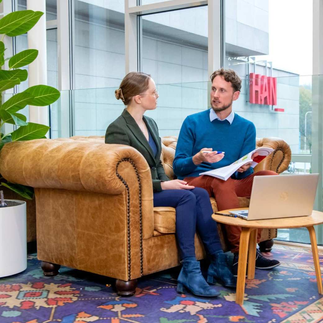 Twee mensen in gesprek op een bank met een plant ernaast, met een laptop op een kleine ronde tafel voor ze